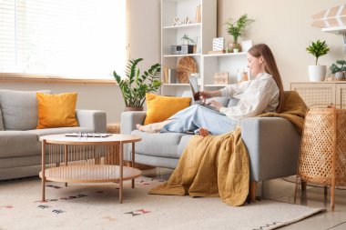 Dizüstü bilgisayar kullanan ve oturma odasındaki kanepede dinlenen güzel genç bir kadın.