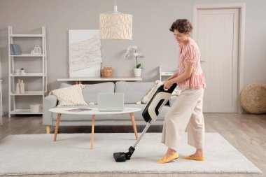 Beautiful young woman cleaning carpet with cordless stick vacuum cleaner in living room