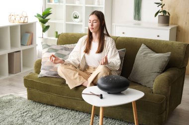 Genç bir kadın evinde glukofonla kanepede meditasyon yapıyor.