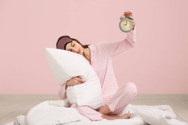 Pembe arka planda battaniyeli ve yastıklı genç bir kadın çalar saat tutuyor.