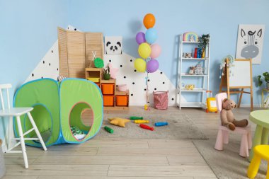 Anaokulundaki modern oyun odasının içinde oyun çadırı, raf ünitesi ve halı üzerinde şekerlemeler var.