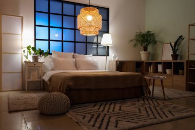 Yatak odasının içinde rahat bir yatak odası, komodin ve parlayan lambalar.