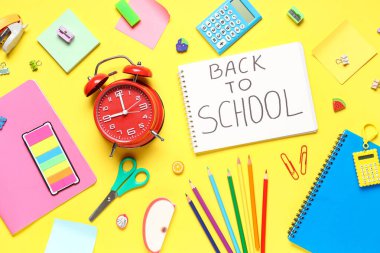 Alarm saati, okula geri yazılan not defteri ve sarı arkaplanda farklı kırtasiye malzemesi