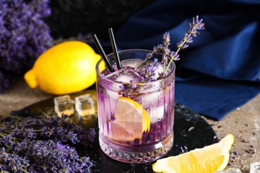 Bir bardak taze lavantalı limonata, çiçekler ve limonlar gri masada.