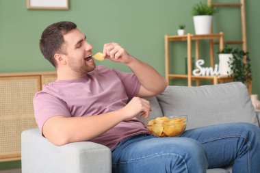 Evdeki kanepede oturan bir kase patates cipsiyle yakışıklı, mutlu genç adam.