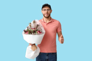 Elinde bir buket güzel çiçekle genç teslimatçı mavi arka planda baş parmak hareketi gösteriyor.