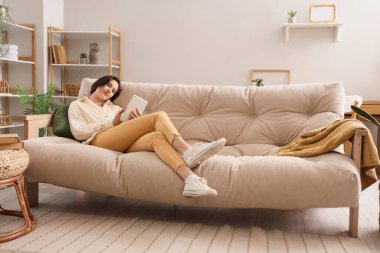 Oturma odasındaki kanepede tablet kullanan genç kadın.