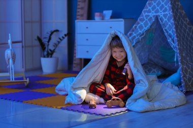 Battaniyenin altında küçük şirin bir çocuk. Parlak el feneri ve karanlık odada oyuncak arabaları var.