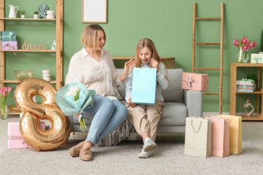 Elinde lale buketi olan güzel bir genç kadın ve elinde kağıt torba ve sekizinci figür balonuyla oturma odasındaki kanepede oturan sevimli küçük kızı.