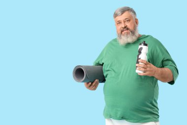Aşırı kilolu, yoga minderli, mavi arka planda bir şişe suyu olan olgun bir adam. Kilo verme konsepti