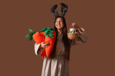 Paskalya tavşanı kulaklı genç bir kadın elinde peluş havuç ve sepetle kahverengi arka planda Paskalya yumurtası tutuyor.