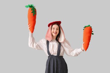 Tavşan kulaklı şık bir kadın, gülümsüyor ve gri arka planda havuç oyuncakları tutuyor. Paskalya kutlaması