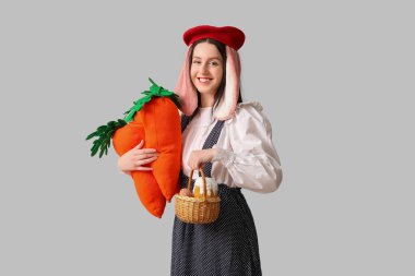 Tavşan kulaklı şık bir kadın, gülümsüyor ve gri arka planda havuç oyuncakları tutuyor. Paskalya kutlaması