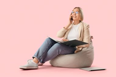 Ofis dosyaları olan olgun bir iş kadını fasulye çuvalında oturuyor ve telefonla pembe arka planda konuşuyor.