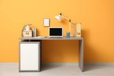 Turuncu duvarın yanında dizüstü bilgisayarı, kırtasiyesi ve lambası olan modern işyeri