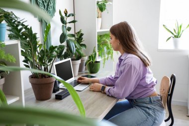 Young woman working with laptop at table and plants in office
