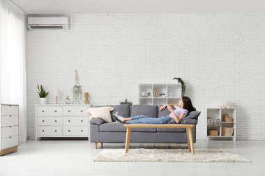 Klima kumandalı genç bir kadın evdeki kanepede uzanıyor.