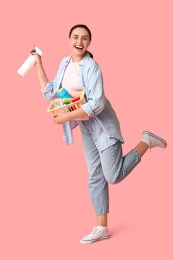 Pembe arka planda farklı temizlik malzemeleriyle dolu sepetli güzel, genç bir kadın.