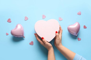 Kırmızı manikürlü kadın elleri, mavi arka planda hediye kutusu ve dekor. Sevgililer Günü kutlaması