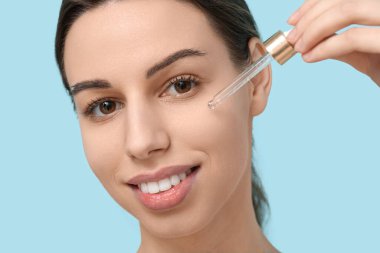 Pipetli güzel genç bir kadın mavi arka planda yüz serumu uyguluyor, yakın plan.