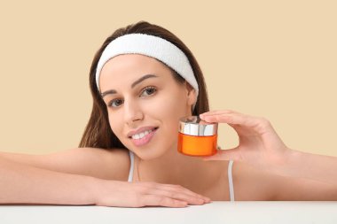 Masanın yanında bej arka planda yüz kremi kavanozu olan güzel genç bir kadın.