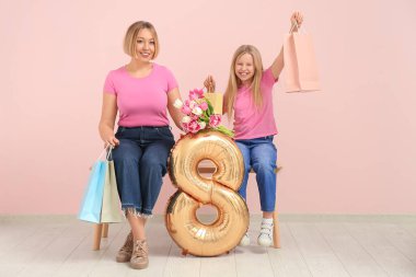 Elinde bir buket lale tutan güzel genç bir kadın ve elinde kese kâğıdı torbaları ve 8 beden şeklinde hava balonu olan sevimli küçük kızı pembe duvarın yanındaki bankta oturuyorlar.