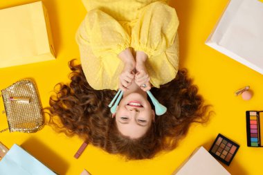 Beautiful young woman with different decorative cosmetics and shopping bags on yellow background