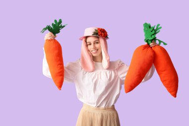 Stylish woman with bunny ears smiling and holding carrot toys on lilac background. Easter celebration