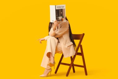 Güzel genç bir kadın dergi okuyor ve sarı arka planda sandalyede oturuyor.