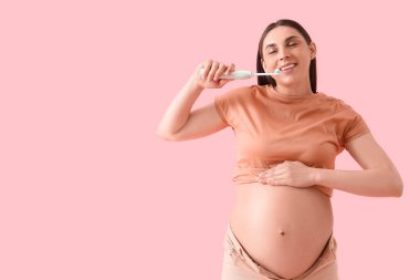 Genç hamile bir kadın pembe arka planda elektrikli diş fırçasıyla dişlerini fırçalıyor.