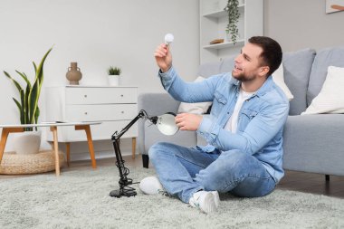 Genç adam oturma odasında masa lambasında ampul değiştiriyor.