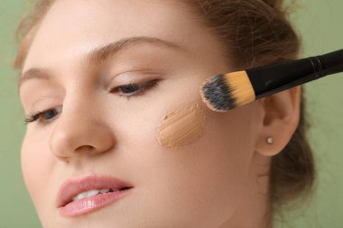 Yüzünde temeli olan güzel genç bir kadın yeşil arka planda makyaj fırçası tutuyor, yakın plan.