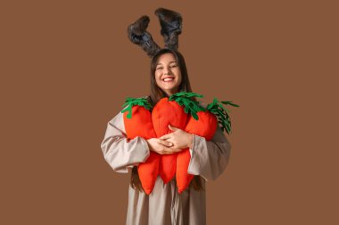 Paskalya tavşanı kulaklı genç mutlu kadın kahverengi arka planda peluş havuç tutuyor.