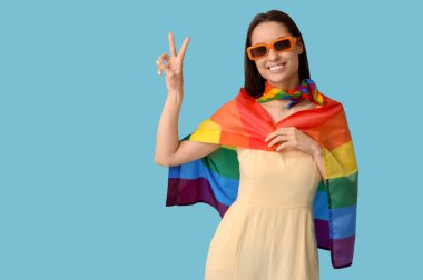 LGBT bayrağı taşıyan, güneş gözlüklü genç bir kadın mavi arka planda zafer jesti gösteriyor.