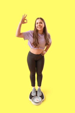 Spor giyimli güzel bir kadın terazide duruyor ve sarı arka planda sorun olmadığını gösteriyor. Kilo verme konsepti
