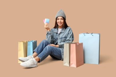 Şık kot elbiseli, şık Asyalı bir kadın alışveriş torbaları ve kahverengi arka planda kredi kartları var.