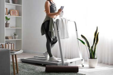 Evde koşu bandında cep telefonu ve su şişesi eğitimi alan sportif bir kadın.