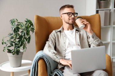 Gözlüklü, dizüstü bilgisayarlı, evde kahve içen genç sakallı bir adam.