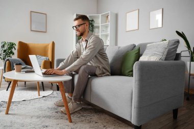 Gözlüklü genç adam evdeki masada dizüstü bilgisayar kullanıyor.