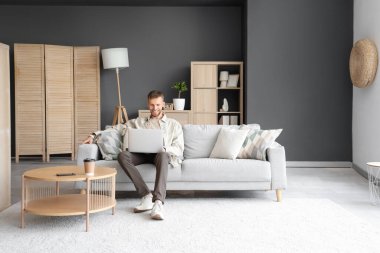 Evdeki kanepede dizüstü bilgisayar kullanan yakışıklı adam.