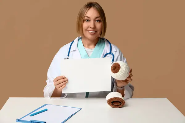 stock image Female doctor with knitted model of breast and blank paper sheet sitting at table on brown background. Breast feeding concept