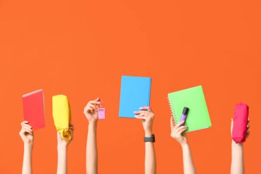 Turuncu arka plan üzerine birçok kalem, kırtasiye ve not defteri olan eller