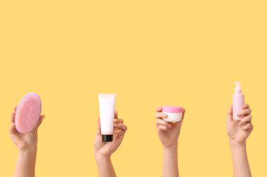 Farklı kozmetik ürünlere sahip kadın elleri ve sarı arka planda saç fırçası