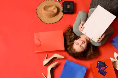 Kredi kartları, şık topuklu ayakkabılar ve kırmızı arka planda alışveriş torbaları olan güzel genç bir kadın.