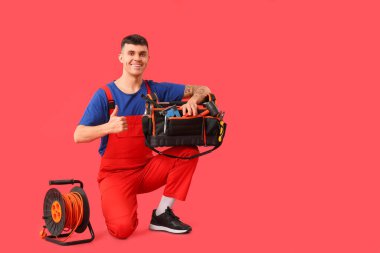 Genişletilmiş kablo makaralı ve alet çantalı genç elektrikçi kırmızı arka planda baş parmağını gösteriyor.