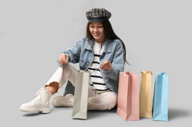 Gri arka planda oturan şık kot elbiseli, alışveriş çantalı, şık Asyalı bir kadın.