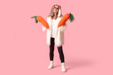 Pembe arka planda havuçları olan tavşan kulaklı güzel bir kadın. Paskalya kutlaması