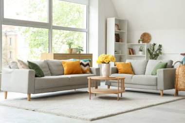 Aydınlık oturma odasının içinde gri kanepeler ve ahşap sehpanın üzerinde sarı narsisli çiçekler.