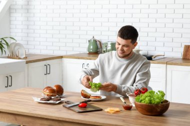 Genç adam, mutfakta lezzetli bir burger için çöreğe marul koyuyor.