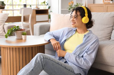 Kulaklıklı genç Afro-Amerikalı kadın evdeki kanepenin yanında oturuyor.